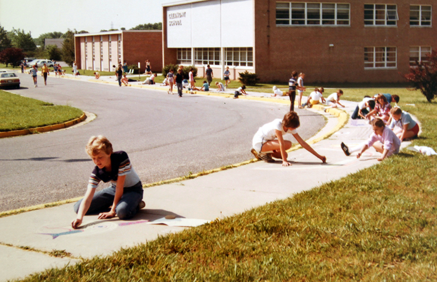 Color photograph of the front exterior of Clermont Elementary School taken in 1981. Several students are sitting on the sidewalk in front of the school coloring the sidewalk with chalk.